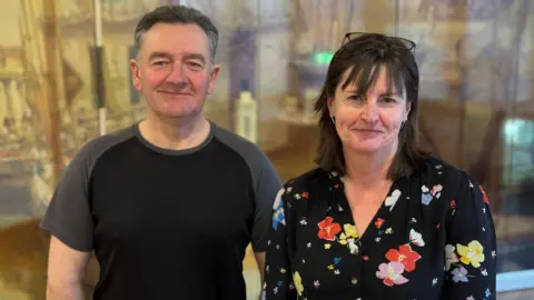 Rob Middleton, who has short greying hair and it wearing a grey and black T-shirt, standing next to Allison Fox, who has shoulder length dark brown hair and is wearing a black blouse with colourful flowers on it and reading glasses on her head.