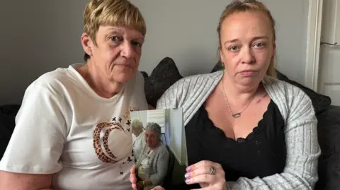Karen Day on the left wears a white shirt with a pattern on it, while sitting next to her daughter Leanne Day, who is wearing a black shirt and a grey cardigan and holding up a photograph of their mother and grandmother, Joyce Sinclair. 