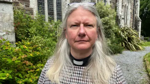  Reverend Claire Reynolds stood outside All Saints Church in Okehampton on Sunday afternoon. She has grey hair and has glasses on her head. 
