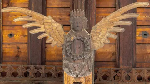 A wooden angel on the roof of St Wendreda's, March. The angel has a crown and its wings are extended in flight. It is holding a wreath and behind it can be seen the roof beams and planks