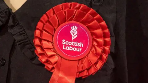 A bright red Scottish Labour rosette with white writing and drawing of a thistle in the middle. It is being worn on the chest of a woman wearing a black jumper and is in close-up.