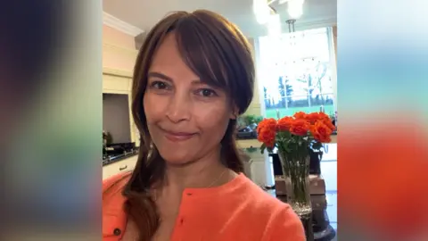 Ratula Chakraborty Ratula Chakraborty looking directly at the camera and smiling. She is wearing a bright orange cardigan and a bunch of red flowers in a vase are on a table behind her.