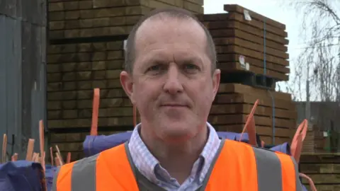 A head and shoulders shot of Simon Pritchard standing in a building merchants' yard. Wooden pallets are stacked behind him. He has short brown hair and is wearing a light-coloured checked shirt, a grey V-neck sweater and an orange hi-vis vest.