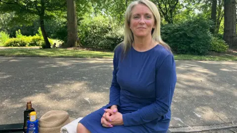 A woman wearing a blue dress s sat in a park. She has long blonde hair and is looking at the camera and smiling. Next to hear are two sun screen products and a straw hat. 