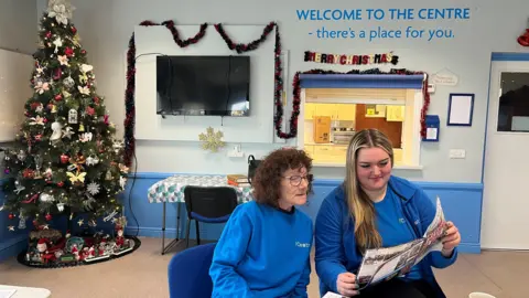 Sharon Barnes and Rebecca Woods reading a copy of Maryport Matters together. They are sat inside the community centre and there are Christmas decorations and a tree in the background.