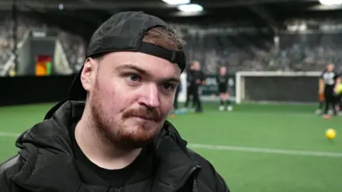 Davison stands on an indoor football pitch. He is wearing a black padded jacket and a black baseball cap worn backwards. The background features a goalpost and several people on the pitch, some wearing dark sportswear and others in bright training bibs. A yellow football is visible on the artificial turf.