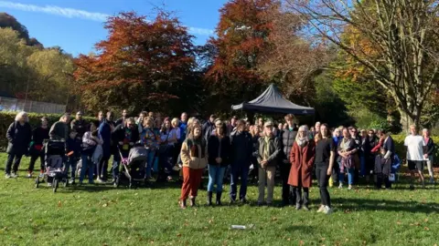 About 100 people stand together in a park with brown autumn leaves behind