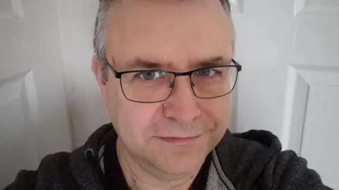 Paul Meadows A selfie of Paul Meadows, who is looking up at the camera. He has black steel-rimmed glasses and is wearing a dark grey hoodie. He has short grey hair.