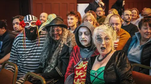 A group of people are sat in an row as part of an audience in full Halloween costumes including a witch and a knight. They are posing for the camera. 