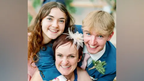 Close-up of three people grouped closely together indoors. In the foreground, one person with short dark hair wears a light-coloured outfit and a decorative white feather hair accessory, along with bracelets and rings. Two younger-looking individuals lean in from either side, both wearing blue suits; one has long brown hair parted in the middle, and the other has short light blond hair and a boutonniere pinned to the suit jacket. All three are positioned tightly together with arms wrapped around each other, against a softly blurred background with greenery."