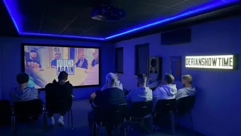 Derian House A view from behind bereaved dads watching Voice Again documentary on a large TV screen inside Derian House Children Hospice's in-house cinema.