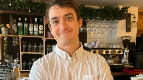 A brown haired man in a striped shirt with a bar behind him.