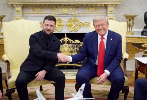 US President Donald J Trump, right, wearing a blue suit, red tie and white shirt, smiles as he shakes hands with President of Ukraine Volodymyr Zelensky, on left, wearing a black suit and with a closed mouth, pleasant expression, in the Oval Office of the White House in Washington DC.