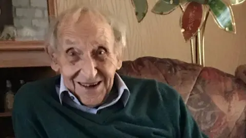 Facebook/Paul Turner Roy Burr, an elderly white man, is smiling at the camera. He is sitting in an armchair, and wears an open-neck blue shirt, and dark green v-necked knitted jumper.