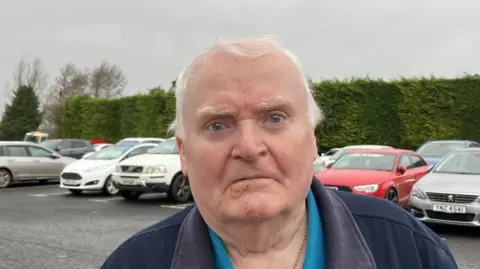 Shows a man with grey hair standing in a car park with cars and a hedge in the background