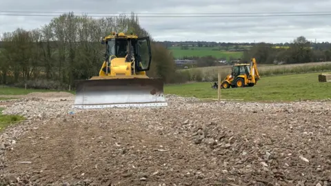 A yellow bulldozer faces the camera with its silver scraper at the front. It sits on a large area of soil that is amid a grassy field, and the soil appears to have been recently turned over. There are trees and fields in the background.