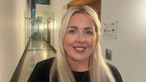 A girl with long blonde hair and blue eyes. She is wearing a black, long sleeve top and a gold necklace. She is standing in a corridor and there is glass door in the background. 