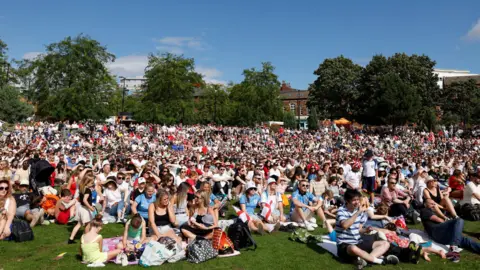 Several thousand people are seated on the grass on Devonshire Green facing centre-left of the camera, looking at a large screen. 
