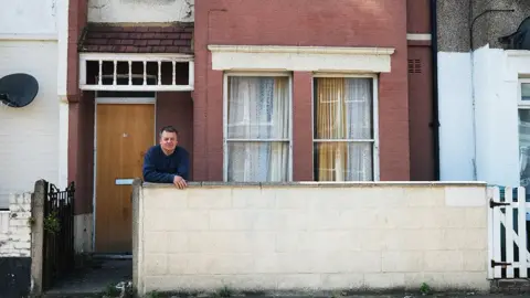 Joe Dixey/BBC Roye Chapman is leaning on a low wall in front of a red brick house with two windows covered by lace curtains and a wooden door. The house has a small front yard with some grass and plants. Roye is wearing a blue long-sleeve shirt