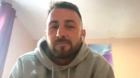 A man with short brown hair and a beard is wearing grey hoodie as he takes a video call from a hotel room
