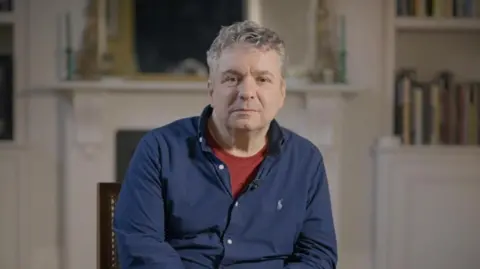 BBC/Adam Walker Dr Ittai Gradel looking to camera in the centre of the image. In blue shirt and maroon top underneath. Behind him books and fire place