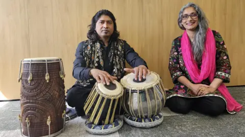 Aisha Iqbal/BBC A man and woman seated on the floor, both wearing traditional South Asian clothing. In front of them are a pair of tabla drums and a dhol drum. The man is playing the tabla drums.