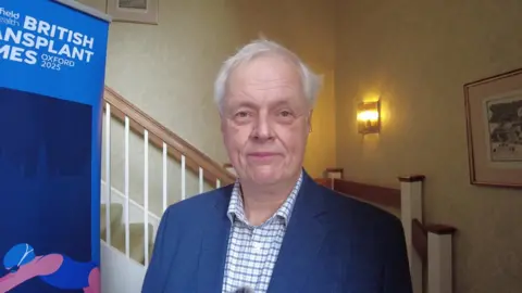 Dr Paul Harden has short grey hair and is wearing a blue blazer over a blue and white chequed shirt. Behind him is a posh looking staircase.