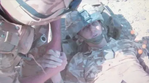 Johno Lee Two soldiers in desert camo army gear. One solider who's face is not in shot is holding the hand of a another solider Johno Lee lying on the floor who has been injured in IED blast. His head is resting on his backpack.