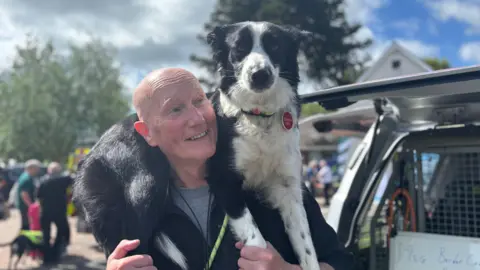 A man is standing with a dog over his shoulders. The man is bald, and smiling widely while looking at the dog. The black and white dog, has a red collar on and is looking at the camera. Behind them is an off-road vehicle and people mingling about. It's a bright sunny day. 