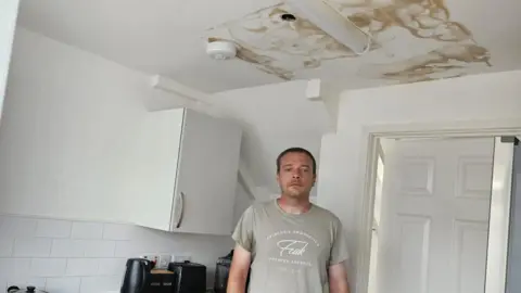 David O'Reilly is stood in his kitchen with a large damp patch on the ceiling with a hole in it. He looks sad in a grey t-shirt.