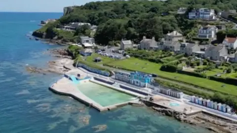 A tidal swimming pool with beach huts and houses behind it.