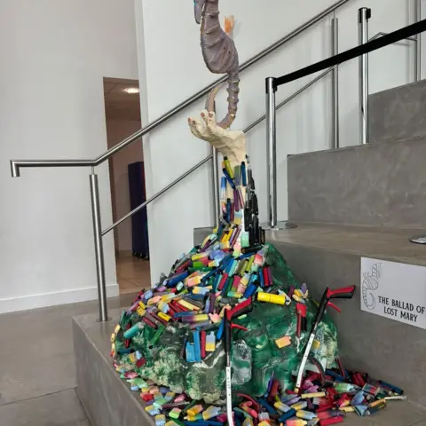 Clean Our Patch CIC A sculpture designed using 4,500 disposable vapes collected by volunteers. The sculpture also has a grey seahorse on top and is positioned at the bottom of a flight of stairs.