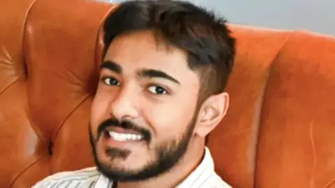 Anugrah Abraham, a man in his early 20s with black hair and thick stubble. His is smiling at the camera while sat on a brown leather sofa.