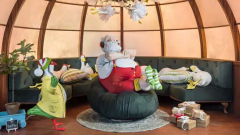 Andrew Meredith Santa in red and white sits on a beanbag. Next to him is Foggy. Behind him on a large sofa are several geese laying down. 