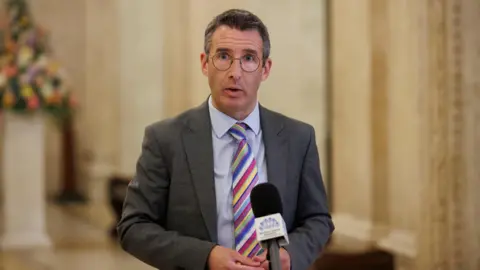 PA Media Minister Andrew Muir standing in front of a microphone. He is wearing a grey suit, blue shirt and stripy multi-coloured tie. He is also wearing glasses. The background of the image is blurred.
