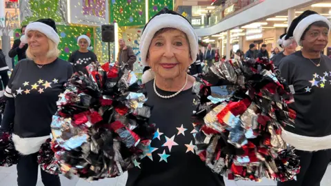 Lily-May Symonds/BBC A close-up of a performer in a shopping centre holding two large pom-poms in red, black, silver, and white. The person is wearing a black sweater with colourful star designs across the chest and a festive hat with white trim. 