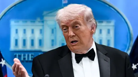 Headshot of Trump who speaks while holding one palm out in front of him gesturing. He stands in front of a blue background white has a white illustration of the White House on it