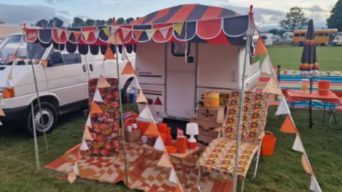 A white caravan is on a pitch of grass with a blue and red awning. Brown and yellow floral chairs are outside the caravan and an orange rug is laid on the grass.