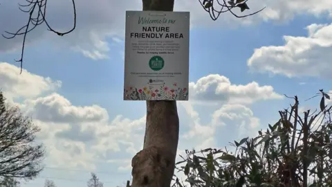 Bradford Council A white sign which reads "welcome to our nature friendly area" attached to a tree trunk with sky and clouds in the background.
