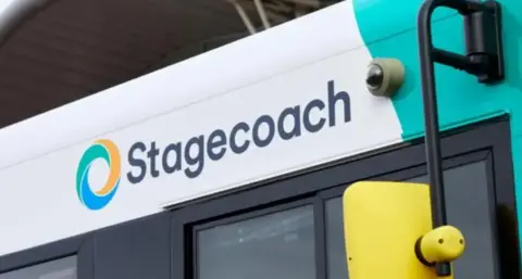 A Stagecoach bus pictured from the side. The driver window is visible, as is a yellow wing mirror and the Stagecoach logo, which is a circle made up of three crescent moon shapes in yellow, green and blue.