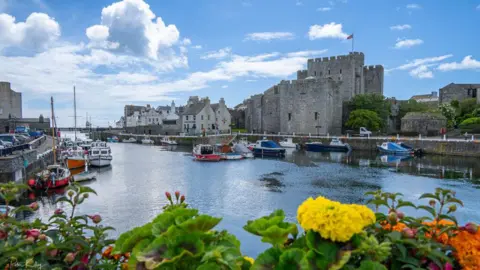 Castletown Harbour on a bright day. There are boats moored in the inner harbour and Castle Rushen can be seen on the right-hand side. There are colourful flowers in the foreground and blue sky with fluffy white clouds above.
