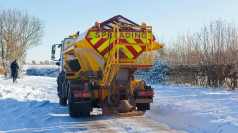 A gritting lorry that says spreading on the back driving down an icy narrow road between snow on banks 