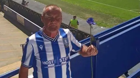 South Yorkshire Police A man with shaved hair wearing a Sheffield Wednesday shirt. He is standing a football ground and holding aloft a blue flower with a long stalk.