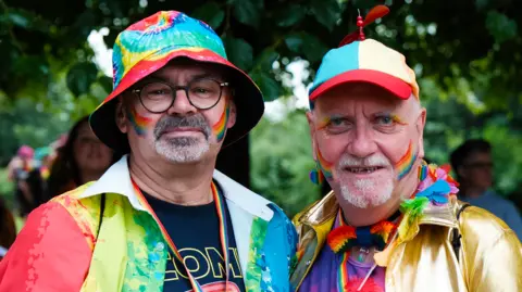 Two Pride parade attendees pose for a photograph outdoors, both wearing colourful clothing and rainbow face paint. One wears a rainbow-patterned bucket hat, while the other wears a metallic gold jacket, rainbow accessories and a multicoloured cap.