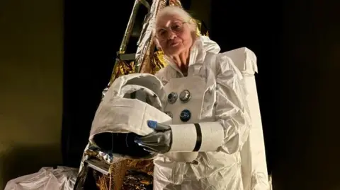 An older woman, with short grey hair, wearing a white spacesuit and holding a space helmet. 