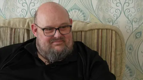 Tom Lawson looking to the right of the camera with pursed lips. He is mostly bald with bits of brown hair on the side of his head and a brown and grey beard. He is wearing black, rectangular glasses and a black top. He is sitting on a beige sofa which has stripes down it. Behind him there is blue and white patterned wallpaper. 