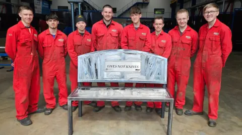 Eight apprentices, wearing identical red boiler suits, stand behind a metal bench made out of surrendered knives. It has a large plaque on the back, reading "LIVES NOT KNIVES".