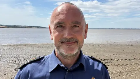 Jake Zuckerman/BBC A man wearing a blue shirt with epilates. He has short hair and a beard and is standing on a shoreline. 
