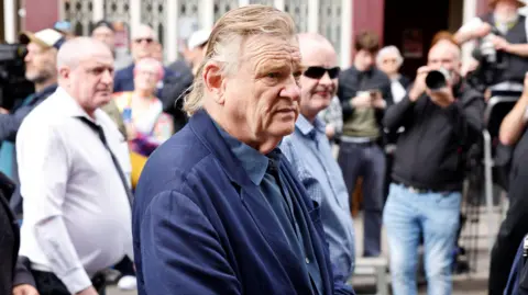 PA Media A man wearing a navy suit and navy shirt hold his hands around the front of his waist. He has medium length red hair as he stands in front of a crowd of people, some of whom have cameras.