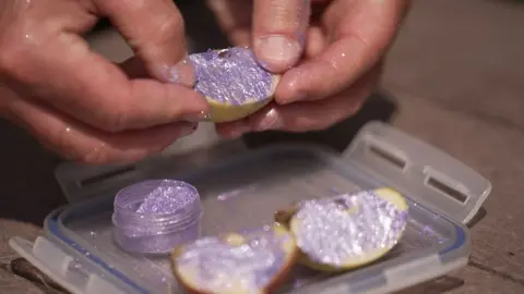 BBC News A man's hands holds an apple which is covered in purple glitter. Underneath the apple is a small glitter container with its cap removed.